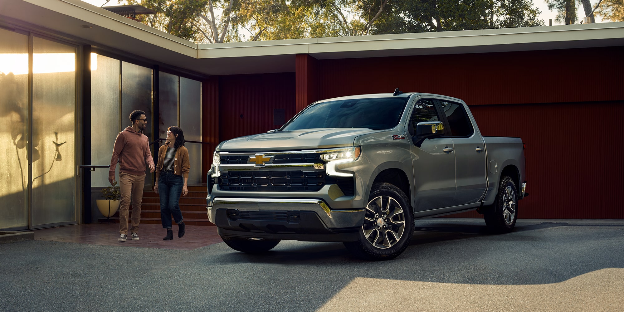 A man and woman walking out to a grey 2025 Chevy Silverado parked in their driveway on a sunny afternoon